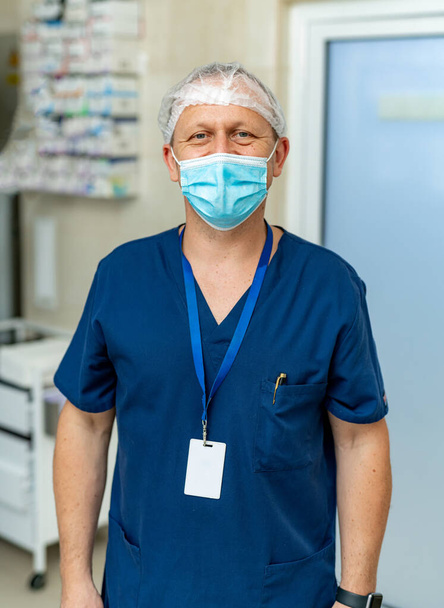 Vertical portrait view of the caucasian male doctor wearing protective mask standing in his cabinet and looking at the camera. Handsome man physician at work in clinic. Healthcare concept - Foto, immagini