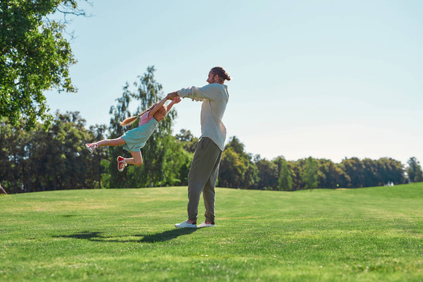Full length shot of young father and daughter having fun, spinning together on grass field in summer park - Photo, image