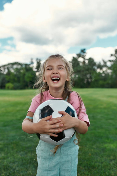 Portrait of joyful little girl holding football and laughing at camera while playing with her parents in park - Foto, afbeelding