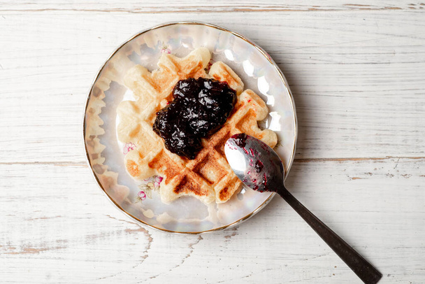 Belgian waffles with blueberry or blackcurrant jam on a plate with a spoon on a white wooden background. - Photo, Image
