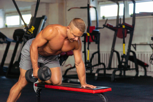 Strongman testépítő gyakorló súlyzóval egy tornateremben.Fiatal jóképű felnőtt férfi csinál egy kar súlyzó sorok padon a modern tornateremben.Másolás tér. - Fotó, kép