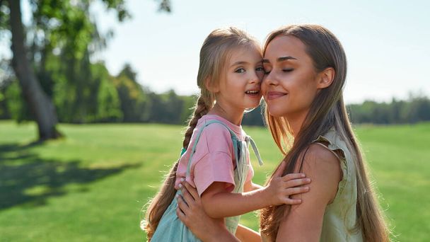 Portrait of happy young mother embracing her cute little daughter, smiling with eyes closed while spending time together in summer park - Foto, Bild