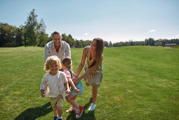 Joyful parents with two little kids playing catch together in green park on a summer day. Happy family enjoying leisure activity - Фото, изображение