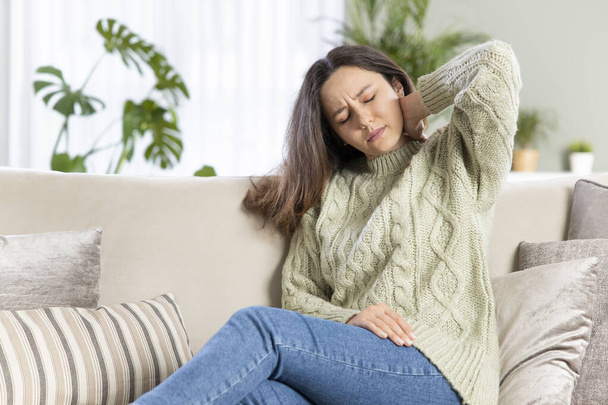 Young woman is sitting at home on her sofa and touching the back of her neck while suffering from neck strain. Neck strain symptoms. - Fotoğraf, Görsel