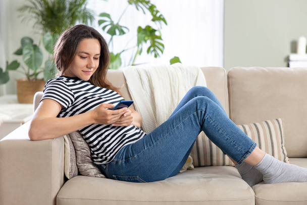 Mladá žena pomocí mobilního telefonu, zatímco sedí pohovka doma  - Fotografie, Obrázek