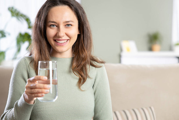 Young woman drinking water sitting on a couch at home. Health benefits of drinking enough water concepts.  - Foto, afbeelding