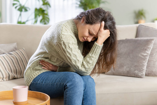 Young woman sitting on the couch at home with a pain in her stomach. Stomach ache, headache, depression concept - Photo, image