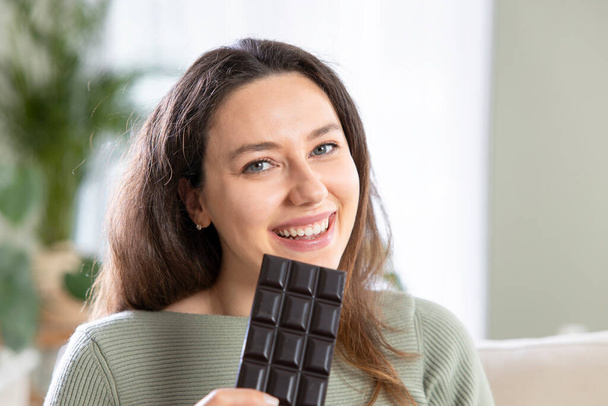 Young woman holding sweet bar of chocolate in living room - Foto, afbeelding