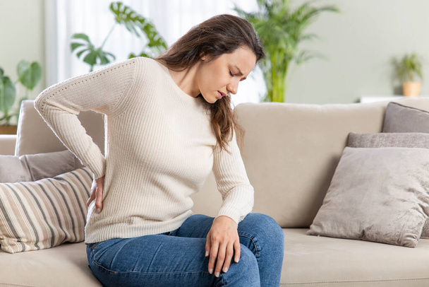 Young woman suffering with back pain, sitting on a couch and holding her lower back with hand. Axial pain, backache - Foto, Bild