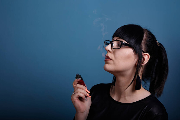 A beautiful girl with dark hair and glasses exquisitely smokes an electronic cigarette on a dark background - Valokuva, kuva