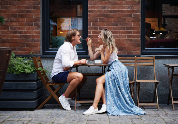 Модная красивая молодая пара, сидящая в кафе и пьющая кофе на улице возле дома. Естественный цвет. Стильная женщина, кормящая ложечника в городской кофейне. Портрет в полный рост - Фото, изображение