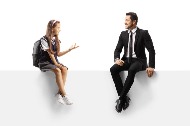 Schoolgirl and a businessman sitting on a blank panel and talking isolated on white background - Foto, Imagem