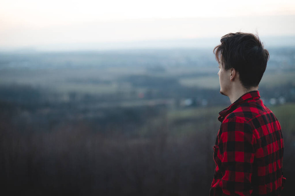 Candid portrait of a man in his 20s dressed in a black and red checked shirt looking into his future, immersed in his thoughts and contemplating. Authentic mood of a young man on an indecisive journey. - Photo, Image