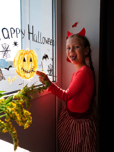 Gyermek az ördögszarvakban sütőtököt fest az ablakra, Halloween-t ünnepli. Kisgyerek rajzol díszíti szoba belső papír denevérek ünneplés ősz nyaralás otthon Kreatív szabadidő lezárása új valóság - Fotó, kép