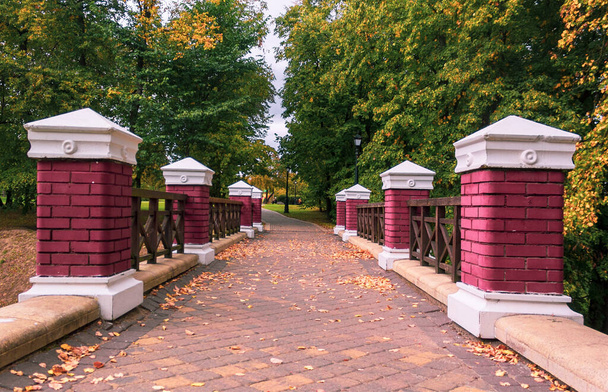 Columnas de ladrillo rojo en la cerca de un puente en un parque de otoño con árboles amarillentos. Hermoso paisaje otoñal en el parque. - Foto, imagen