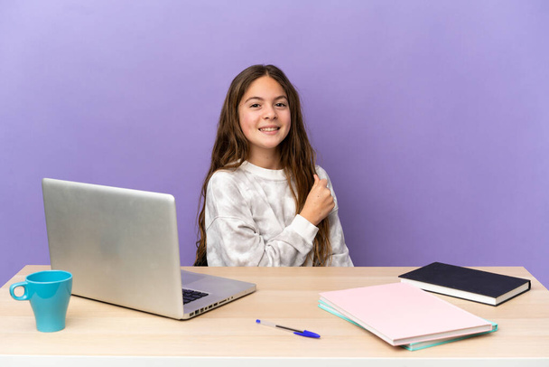 Kleines studentisches Mädchen an einem Arbeitsplatz mit einem Laptop auf violettem Hintergrund, der einen Sieg feiert - Foto, Bild