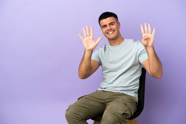 Jongeman zit op een stoel over geïsoleerde achtergrond tellen negen met vingers - Foto, afbeelding
