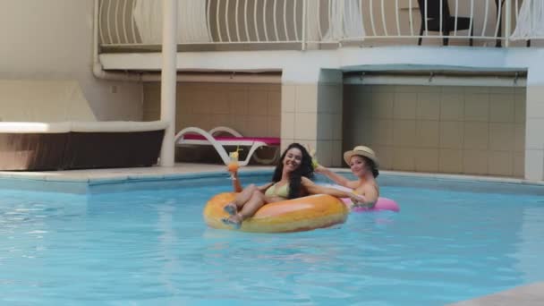 Glücklich lachende Studentinnen, die gemeinsam unbeschwerte Sommerferien genießen. Europäische Blondine und Latina Brünette schwimmen auf Gummiringen und trinken Eissaft-Cocktails und amüsieren sich am sonnigen Wochenende - Filmmaterial, Video