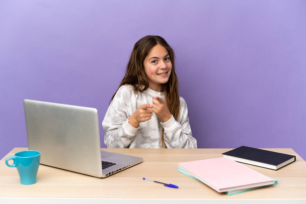 Little student girl in a workplace with a laptop isolated on purple background surprised and pointing front - Foto, Imagem