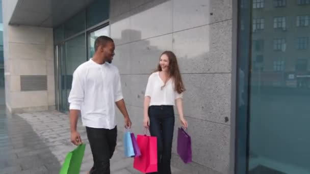 Νεαρό θετικό πολυεθνικό ζευγάρι φεύγει από το κατάστημα μαζί με αγορές περπατώντας στην πόλη σε εξωτερικούς χώρους. Χαρούμενη ποικιλόμορφη αφρο-αμερικανός άνδρας και καυκάσιος γυναίκα με τα πόδια έξω από το εμπορικό κέντρο με τσάντες ψώνια μιλάμε - Πλάνα, βίντεο