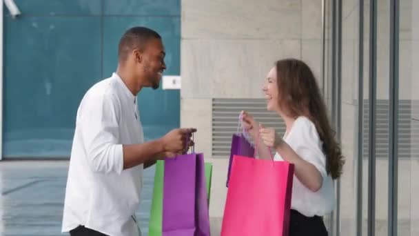 Çeşitli etnik çeşitliliğe sahip mutlu çift, çok ırklı erkek ve kadın alıcılar alışveriş merkezinin yakınında eğleniyorlar. Birlikte dans ediyorlar. Parlak çantalar satın alıyorlar. - Video, Çekim