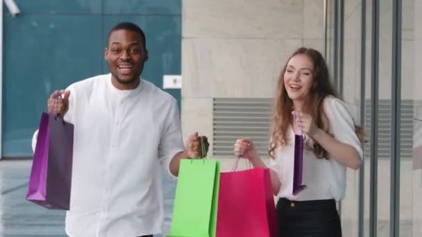 Beautiful happy young multiracial multiethnic couple with shopping bags dance outdoors sing song joyful moving black friday discounts. Cheerful man and woman funny dancing with purchases near store - Footage, Video