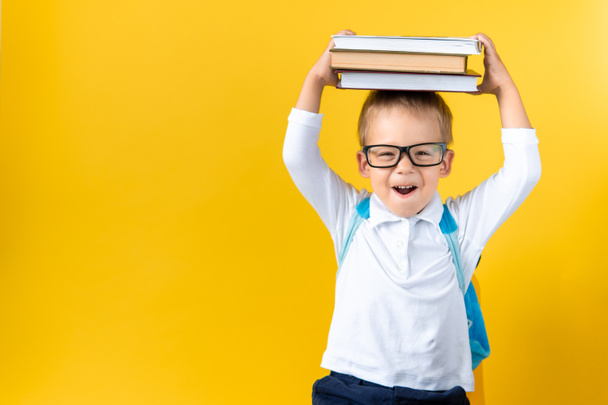 Banner Funny Preschool Child Boy in Glasses with Book on Head and Bag on Yellow Background Copy Space. Happy smiling kid go back to school, kindergarten. Success, motivation, winner, genius concept. - Photo, Image
