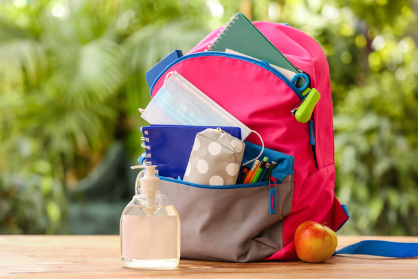 Schulrucksack mit Schreibwaren, medizinischer Maske, Flasche Desinfektionsmittel und Apfel auf dem Tisch im Freien - Foto, Bild