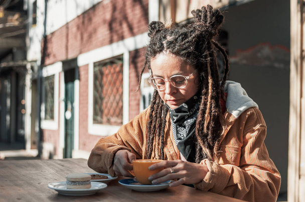 pretty young hispanic latin girl with glasses, black scarf, brown corduroy jacket and dreadlocks enjoys sitting at a table outside a coffee shop and holds a cup of coffee in her hands to drink it. - Foto, Imagem