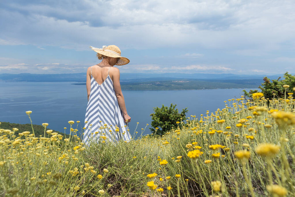 Vue arrière de la jeune femme portant une robe d'été rayée et un chapeau de paille debout dans une super floraison de fleurs sauvages, relaxant tout en profitant d'une belle vue sur la nature de la mer Adriatique, Croatie - Photo, image