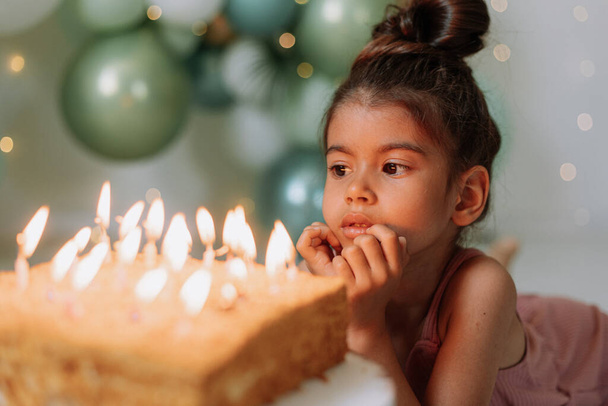 Симпатичная девочка с праздничным тортом дома на фоне воздушных шаров. День рождения ребенка - Фото, изображение
