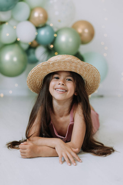Aranyos gyermek lány rózsaszín ruha és szalma kalap ünneplő születésnap, színes lufik háttér - Fotó, kép