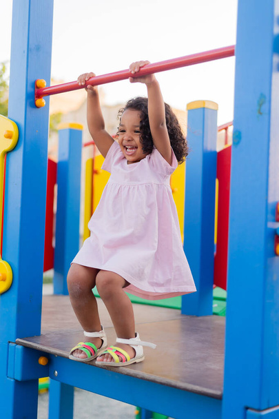 Cute African American little girl with curly hair and pink dress playing at playground - Photo, image