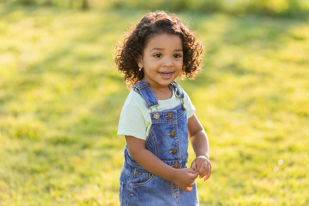 Small dark-skinned girl with curly hair in a denim sundress walks at park on a sunny day. Concept of a healthy lifestyle. Happy childhood - Photo, Image