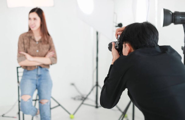 Selective focus on asian male photographer holding camera and taking photo of blur background long hair woman in studio with lighting equipments, tripods and white cutout - Photo, Image