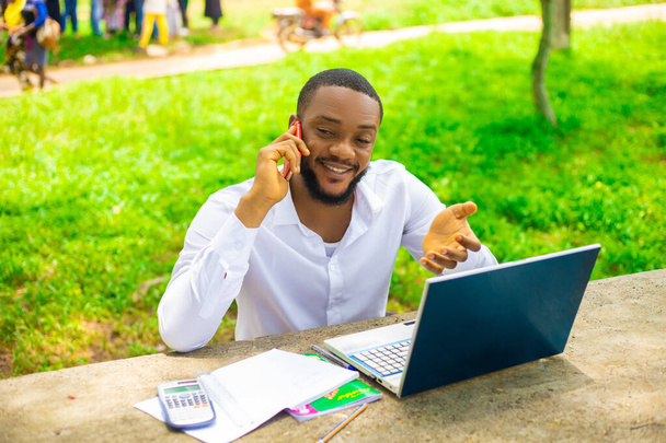 A handsome African male student talking on the phone sitting by the table in the campus park - Photo, Image