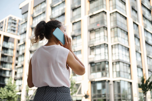 Kentsel yüksek binaların arka planında cep telefonuyla konuşan genç bir Afro-Amerikan, Latin kökenli kadının görüntüsü. İş ve iletişim kavramları - Fotoğraf, Görsel