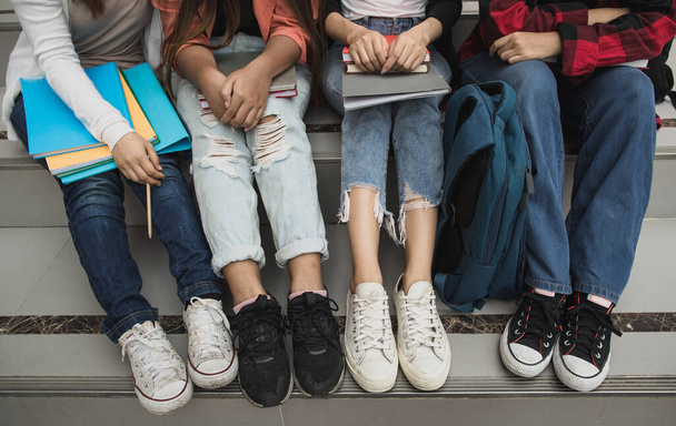 Grupo de cuatro jóvenes estudiantes universitarios piernas y zapatillas de deporte sentados juntos en el campus universitario al aire libre. Concepto para la educación, la amistad y la vida universitaria. - Foto, Imagen