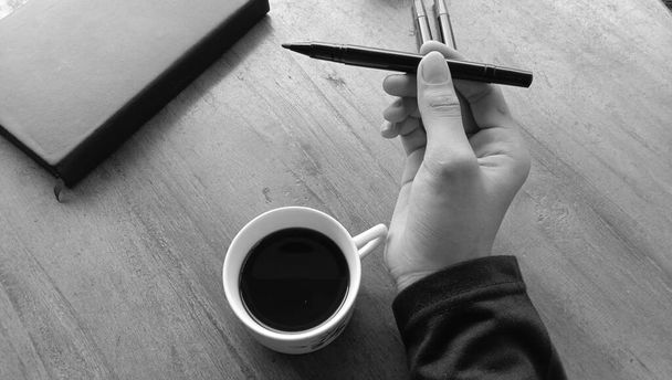 A top grayscale view of a man's hand holding a pen near a cup of coffee - freelance and break concept - Foto, Imagen