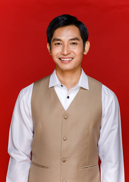 Young attractive Asian man soon to be groom wearing white shirt and beige vest smiling at camera against red background. Concept for pre wedding photography. - Photo, Image