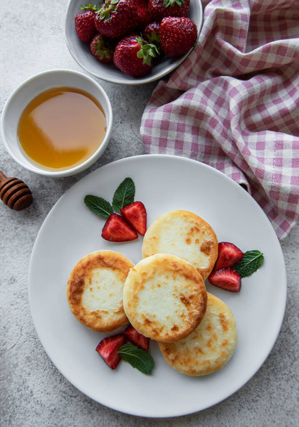カッテージチーズパンケーキ、新鮮なイチゴとセラミックプレート上のリコッタフリッター。健康的でおいしい朝の朝食。灰色のコンクリートの背景. - 写真・画像