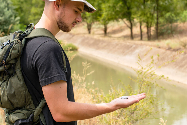 a tourist guy with a hat and a backpack is guided by a compass standing on the river bank. High quality photo - Photo, Image
