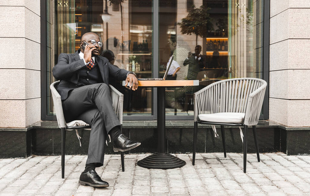 Portrait of a black African American businessman in a suit sitting in a city cafe outdoors and talking on the phone. - Photo, image