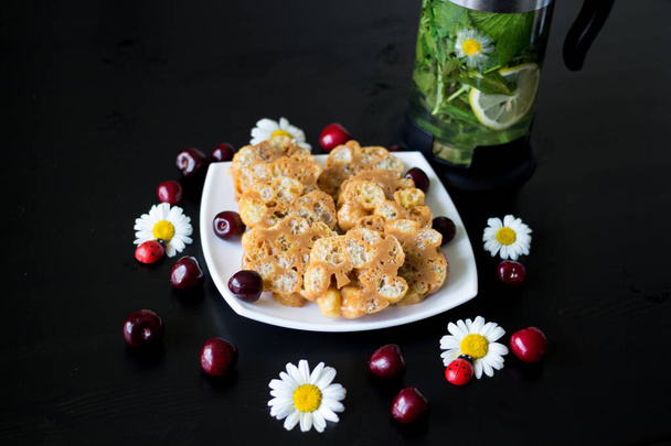 Dessert of toffee and corn sticks on a white square plate, decorated with daisies with ladybirds and cherries. Next to the kettle, which is brewed currant leaf, lemon, mint and chamomile. - Photo, Image