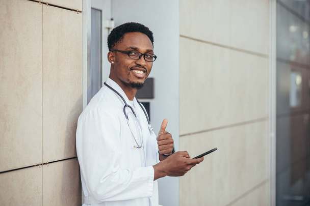 A cheerful man of an African American doctor, near the clinic, in a white medical gown looks at the camera and holds a mobile phone to talk to patients smiling at the camera reports good news - Фото, изображение