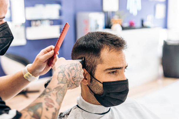 Barber cutting the hair of a client with a mask with a machine - Photo, Image