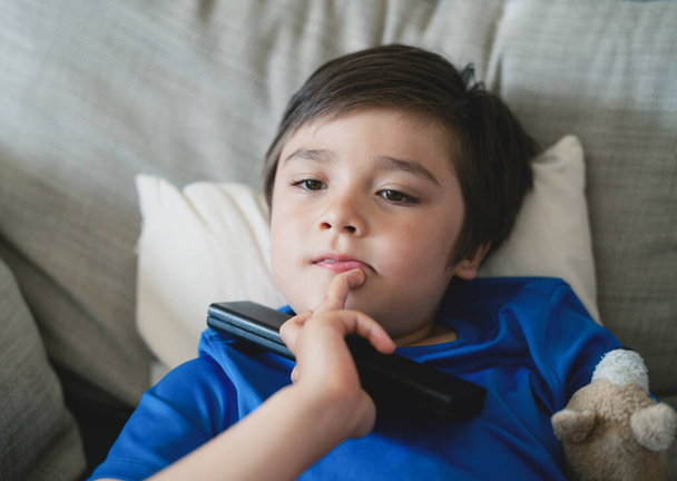 Happy mixed race boy putting finger on his chin while watching TV, Candid shot Youg kid lying on sofa holding remote contro, Positive child relaxing at home on weekend - 写真・画像
