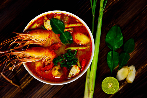 Top view shot of Fresh delicious tasty Tom Yam Kung hot sweet sour spicy Thai shrimp prawn mushrooms and herb soup in white bowl on sack cloth with ingredients spring onion lemon basil on wood table. - Photo, Image