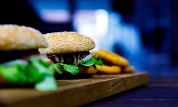 Closeup shot of delicious tasty yummy hamburger made with sesame seed bun fresh cooked meat tomato and lettuce serving with fried onion rings on wood cutting board plate on dark old wooden table. - Фото, изображение