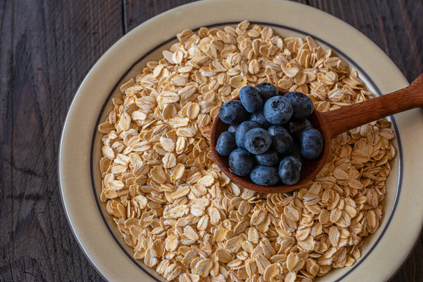 Oatmeal with blueberries. Healthy breakfast ingredients with rolled oats and fresh blueberries served on wooden table. Closeup and table top view - Photo, Image